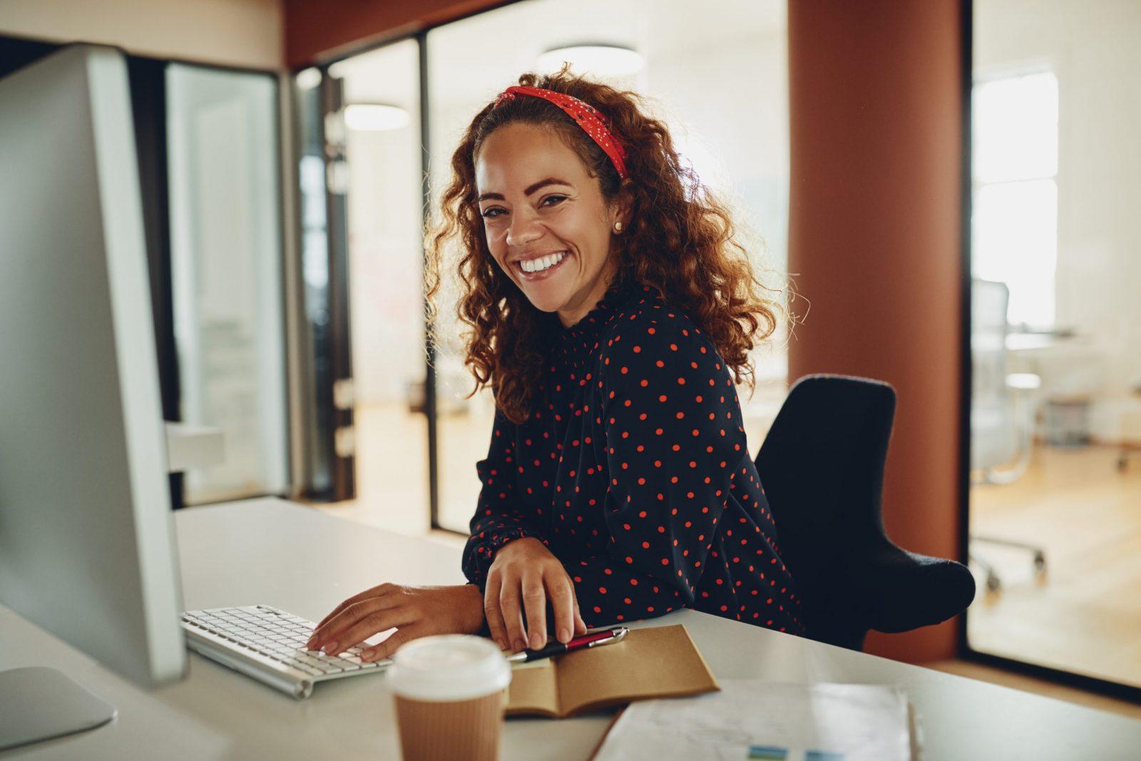 Young businesswoman sitting at her desk and smiling while working on a computer and taking notes